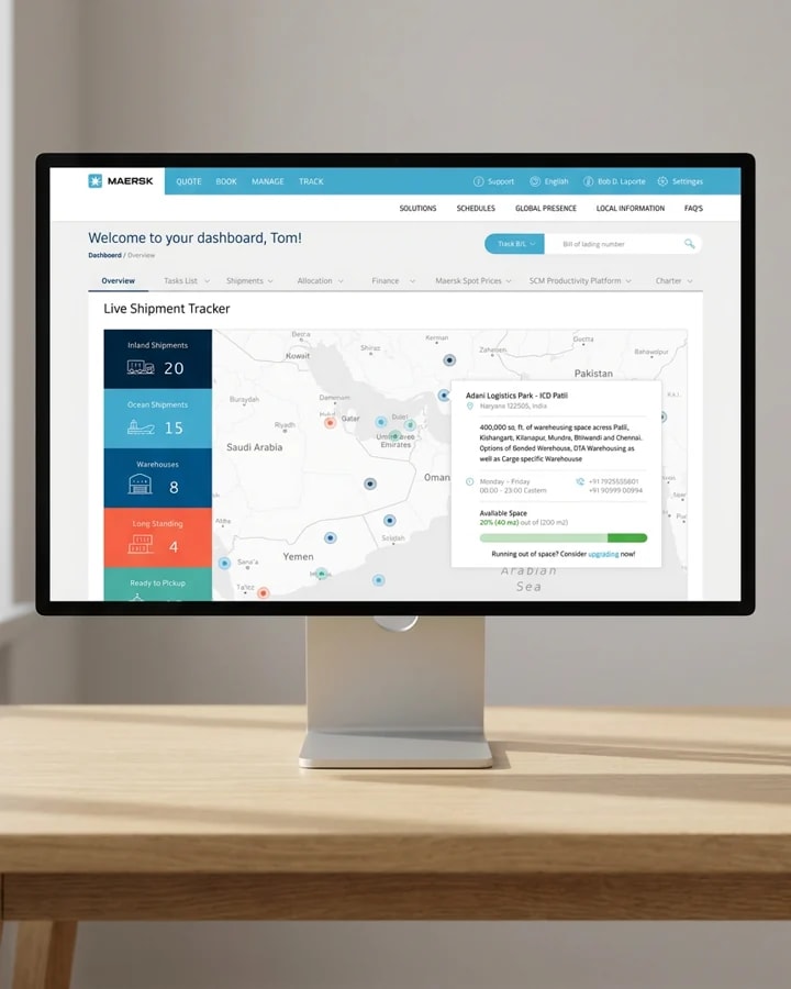 A desktop monitor on an oak desk showing the Maersk Hub portal: a live shipment tracker with counts for inland shipments, ocean shipments and warehouses beside a map of the Gulf.